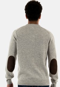 Persoon met krullend haar, die een lichtgrijze trui met donkere, ovale elleboogpatches draagt, gezien vanachter tegen een effen achtergrond.