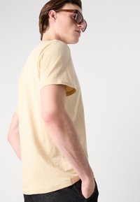 Lys gul bomulds t-shirt med rund hals og korte ærmer, vist fra siden. Mørke jeans og store brune solbriller er også synlige.