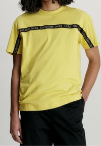 Žluté bavlněné tričko s černým pruhem se značkou "Calvin Klein Jeans" přes hruď a na rukávech. Uvolněný střih s kulatým výstřihem.