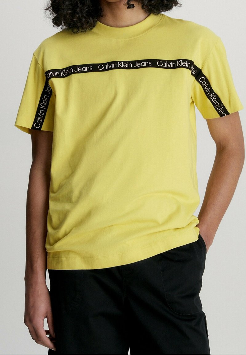 Žluté bavlněné tričko s černým pruhem se značkou "Calvin Klein Jeans" přes hruď a na rukávech. Uvolněný střih s kulatým výstřihem.
