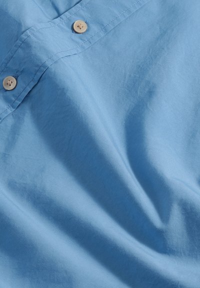 Lichtblauwe stof met een gladde textuur en een subtiele glans, gedetailleerd met beige knopen en een flat-col ontwerp.