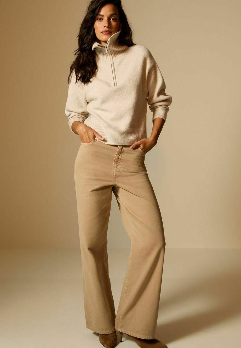 Suéter beige con cremallera y puños acanalados combinado con pantalones de pierna ancha en un tono claro. La modelo posa con las manos en los bolsillos frente a un fondo neutro.