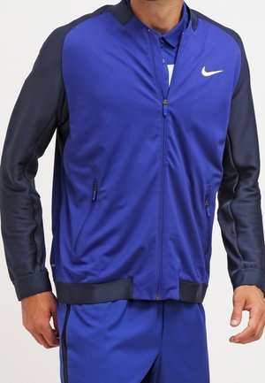 Mężczyzna w niebiesko-czarnej sportowej kurtce zapinanej na zamek z białym logo Nike, w parze z dopasowanymi niebieskimi sportowymi spodniami.