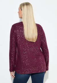 Bordeauxfarbene Langarmbluse mit metallischen Sprenkeln, abgerundetem rückseitigen Saum und weicher Textur, kombiniert mit dunkelblauen Jeans.