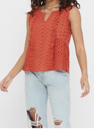 Frau trägt ärmelloses rost-orangefarbenes Top mit Lochmuster und Rüschen sowie hellblaue zerrissene Jeans, steht vor weißem Hintergrund.