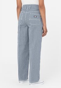 Personne portant un pantalon large bleu et blanc à rayures verticales taille haute avec poches arrière et des baskets blanches, debout devant un fond uni.