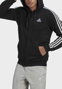 Moletom preto com fecho e capuz, feito de mistura de algodão, apresentando um capuz, bolsos frontais e três riscas brancas nas mangas. Logótipo da Adidas no peito.