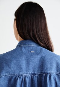 Denim shirt with a gathered back and a metallic tag near the collar. Features a classic collar and a soft, lightweight texture.