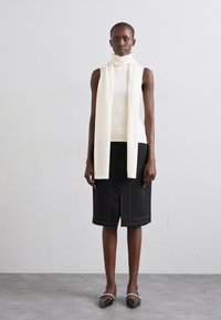 Un haut sans manches blanc à col montant avec une écharpe assortie, associé à une jupe noire longueur genoux avec des coutures contrastantes et une coupe droite.