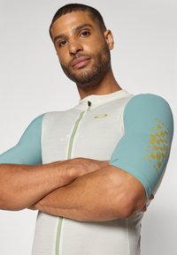 Haut de sport à manches courtes en gris clair et bleu clair, présentant une fermeture éclair sur le devant et des accents à motifs sur les manches. Conçu pour le confort et la performance.