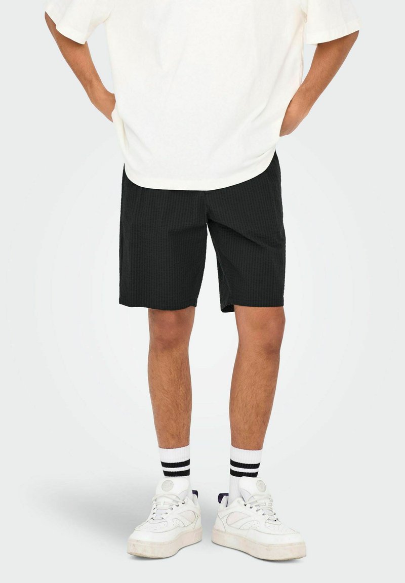 Zwarte shorts met een textuurpatroon, gedragen met een oversized wit t-shirt. Gecombineerd met witte sneakers en zwart-witte gestreepte sokken.