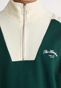 Groene en crèmekleurige ritssweater met een hoge kraag, voorzien van een zilveren rits en een geborduurd logo aan de linkerkant.