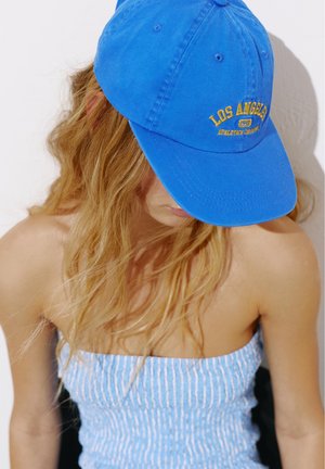 Jeune femme aux longs cheveux blonds porte une casquette de baseball bleue « Los Angeles » et un top rayé bleu et blanc sans bretelles.