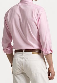 Camicia rosa chiaro a maniche lunghe con polsini arrotolati, dotata di colletto e tessuto morbido. Indossata con jeans bianchi e cintura marrone. Vista da dietro.