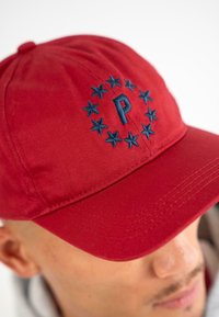 Căciulă roșie din țesătură, cu bor constant, având litera „P” brodată în albastru, înconjurată de stele aranjate într-un model circular.