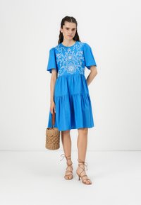 Blue dress with floral embroidery on the bodice, short puff sleeves, and a tiered skirt. Accompanied by a woven handbag and strappy heels.