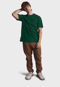 Zelené bavlněné tričko s krátkým rukávem v kombinaci s hnědými kalhotami. Modelka má na sobě bílé tenisky a pózuje s jednou rukou na hlavě.