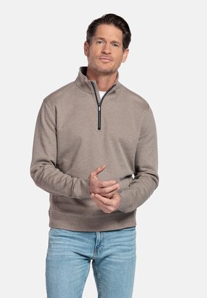 Man draagt een beige pullover met kraag en lichtblauwe spijkerbroek, staand tegen een effen lichte achtergrond, handen samengevouwen voor zich.