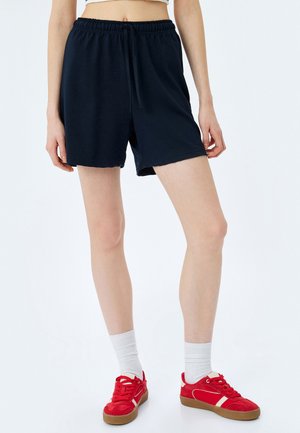 Marineblaue Shorts aus weichem Stoff, mit einem elastischen Bund und einer Kordel zum Verstellen, in lockerer Passform. Getragen mit roten Sneakers und weißen Socken.