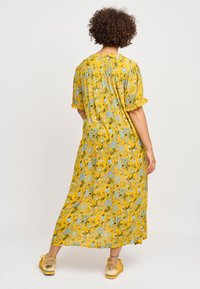 Gul blomstrede maxikjole med korte ærmer, blå og grønne mønstre, tekstureret stof og bølget kant. Parret med gule sko.