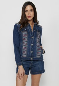 Jeans jakna z izvezenimi sprednjimi paneli, gumbnim zapiranjem in dvema žepoma na prsih. Temno modra barva, strukturiran kroj in dolgi rokavi.