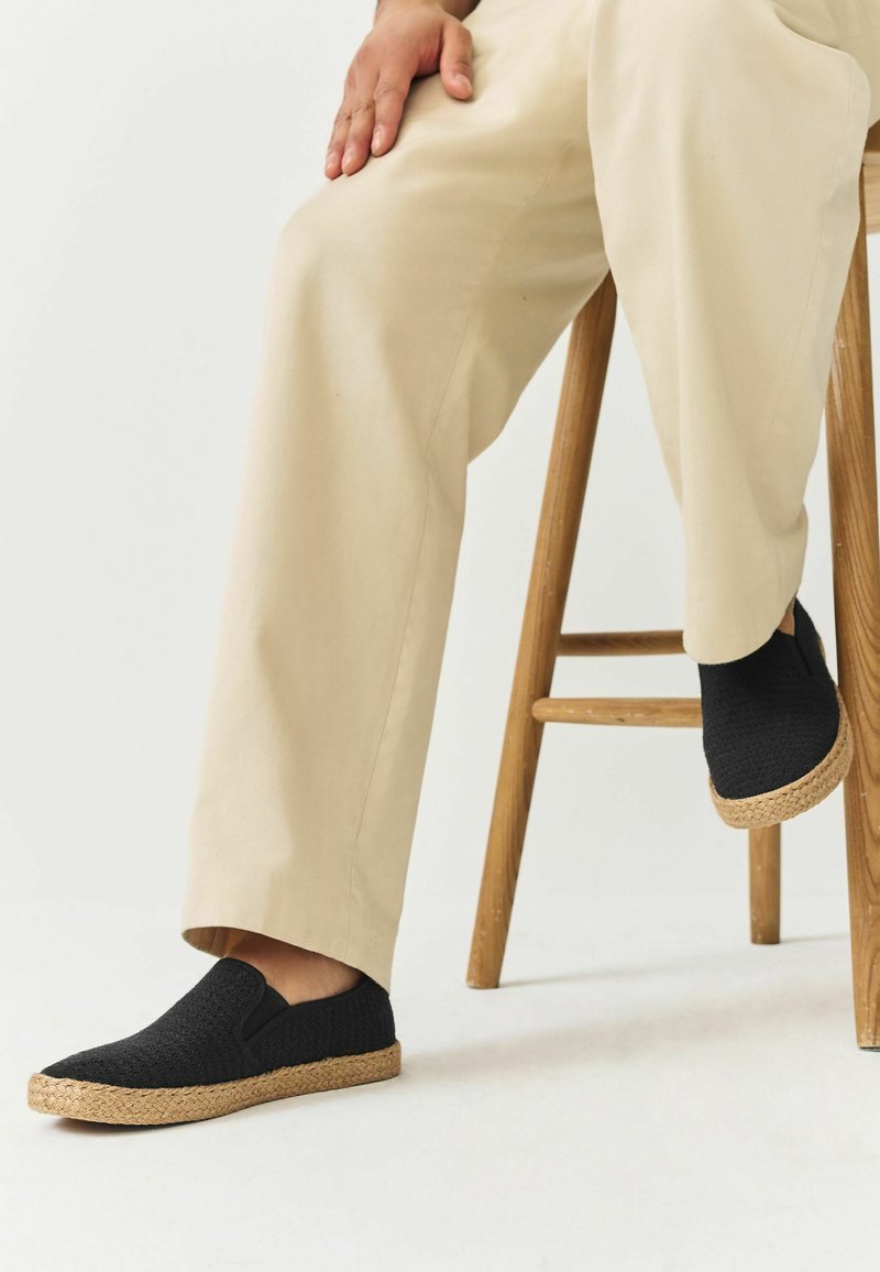 Persona sentada en un taburete de madera, con pantalones beige y zapatos slip-on negros tejidos con suelas de color natural.