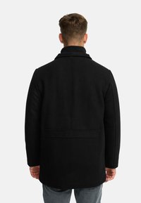 Schwarzer Wollmantel mit hohem Kragen und strukturiertem Schnitt, ausgestattet mit einer Rückennaht und Taschen, kombiniert mit blauen Jeans.