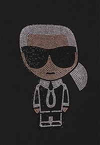 Figure stylisée avec des cheveux blancs en queue de cheval, lunettes de soleil noires et costume noir et blanc composé de petits strass sur tissu noir.