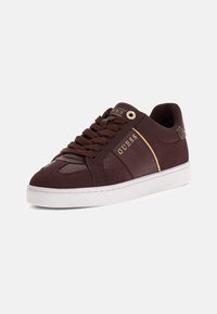Brune ruskinds-sneakers med guldaccenter, hvid sål, snørebåndsdesign og perforerede detaljer på tåen og siden. Brandlogoet er fremhævet.