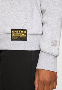 Sudadera gris con puños y dobladillo acanalados, con una etiqueta negra y dorada de G-Star Originals en el frente, usada sobre tela negra.