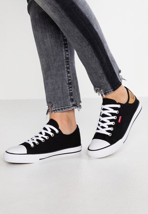 Person iført sorte og hvide lærreds sneakers med hvide snørebånd og mørkegrå jeans med frynset kant, stående på en hvid overflade.