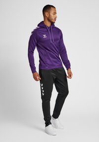 Felpa con cappuccio viola e dettagli neri e logo, abbinata a pantaloni jogger neri con un motivo a chevron bianco lungo il lato, indossata con sneaker bianche.