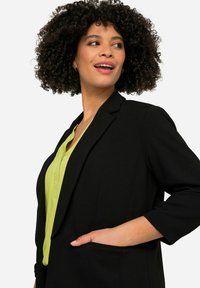 Blaser noir au design ajusté, avec des revers crantés et une poche simple. Associé à un chemisier vert vif, avec un col et des boutons visibles.