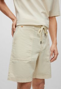 Béžové šortky na stažení se širokými předními kapsami, které nosí osoba s rukama položenýma na bocích.