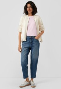 Veste bombardier crème, t-shirt rose clair, jean bleu taille haute et baskets argentées avec des accents beige. Cheveux bouclés et lunettes.