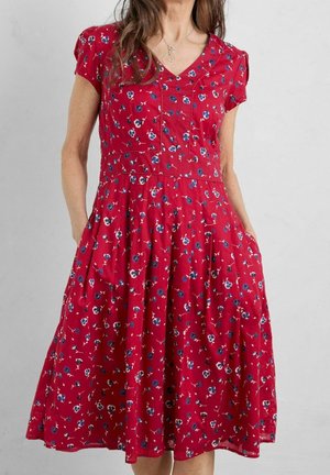 Femme portant une robe rouge à fleurs jusqu'aux genoux, à manches courtes, encolure en V et poches, debout devant un fond gris uni.