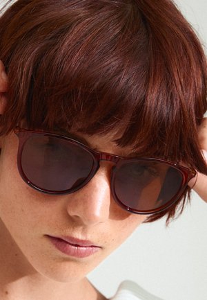 Gafas de sol tintadas en marrón con una montura translúcida de color burdeos, lentes redondeados y detalles sutiles en las varillas, sobre un fondo claro.