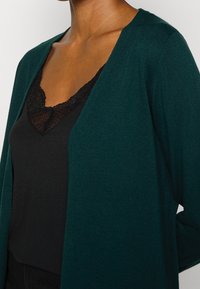 Cardigan de un verde oscuro con una textura suave y un diseño de frente abierto, superpuesto a una blusa de camisón negra con encaje.