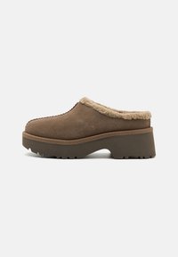 Zoccoli in suede marrone con fodera in pelliccia sintetica, punta arrotondata e una robusta suola piatta e texture. Dettagli di cuciture a contrasto lungo i bordi.