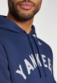 Jūras spēku kapuce, kas rotāta ar baltu izsistām burtiem "YANKEES", ar auklu uz galvas un mīkstu faktūru. Tuvplāns uz kakla daļu un šuvēm.