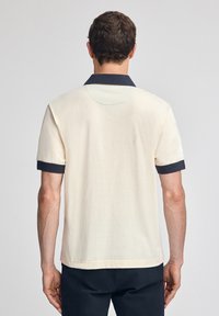 Krämfärgad kortärmad polo skjorta med marinblå krage och manschetter, med en slät textur och öppen nacke.