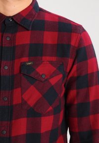 Czerwona i czarna koszula flanelowa w kratę, z kołnierzykiem na guziki, kieszenią na piersi i czarnymi zatrzaskami. Miękka w dotyku o klasycznym wzorze.