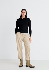 Camisa polo negra de manga larga con cuello, combinada con pantalones beige. El modelo lleva botas de tobillo verde oliva con suelas negras y detalles de botones.