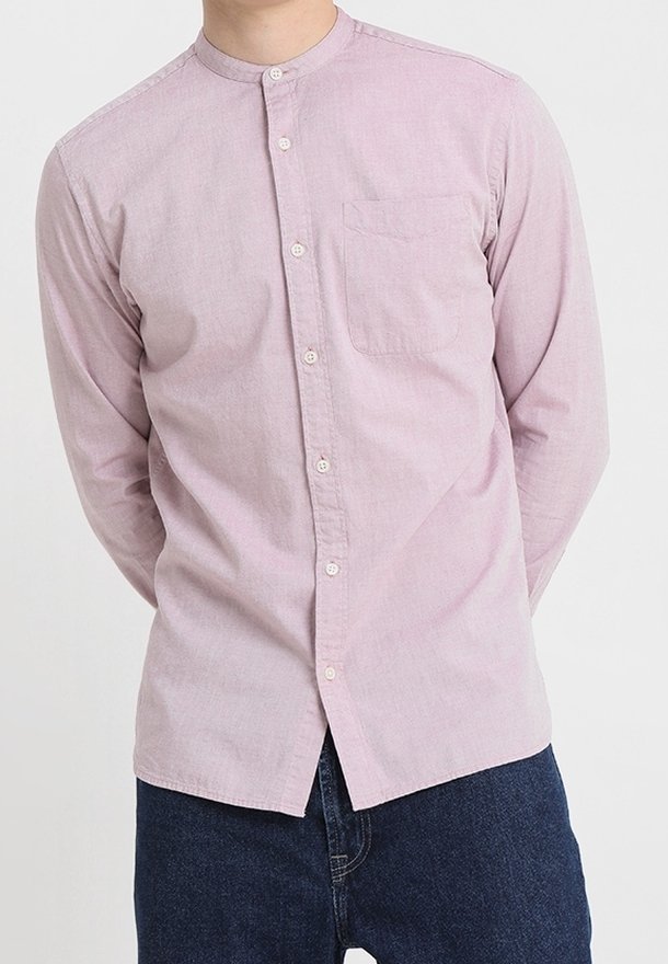 Man wearing light pink button-up shirt with mandarin collar and dark blue jeans, standing with hands behind back against white background.