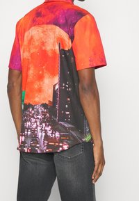 Chemise à manches courtes présentant un graphisme vibrant rouge et violet d'un paysage urbain au clair de lune, avec des détails complexes et une texture lisse.