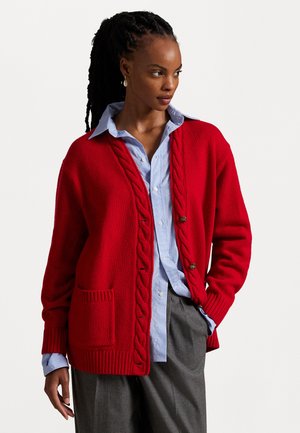 Roter Strickcardigan mit Rippstruktur, Zopfmuster und zwei Knöpfen. Kombiniert mit einem hellblauen Hemd mit Kragen und grauen Hosen.