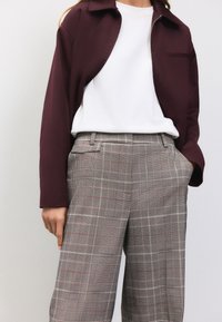 Giacca bordeaux con zip sopra una camicia bianca, abbinata a pantaloni a vita alta grigi a quadri con linee rosse di accento e tasche laterali.
