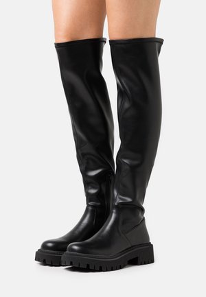 Bottes en cuir noires montant jusqu'aux genoux avec semelles épaisses à relief, portées par une personne debout devant un fond clair uni.