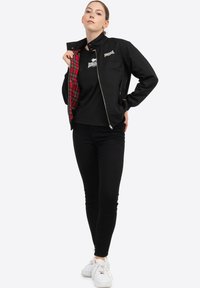 Schwarze Zip-Jacke mit kariertem roten Innenfutter, kombiniert mit einem schwarzen T-Shirt und schwarzen engen Jeans. Das Modell trägt weiße Sneaker.