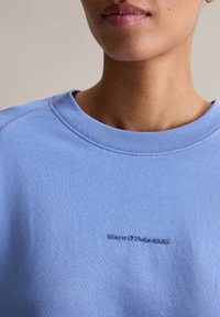 Felpa azzurra chiara con scollo tondo, caratterizzata da rifiniture a coste e un dettagli logo blu scuro sul davanti. Texture liscia e design casual.